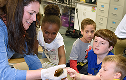 Female educator wearing a glove holds a specimen while four young children gather around, closely observing and engaging with the activity in a classroom setting.