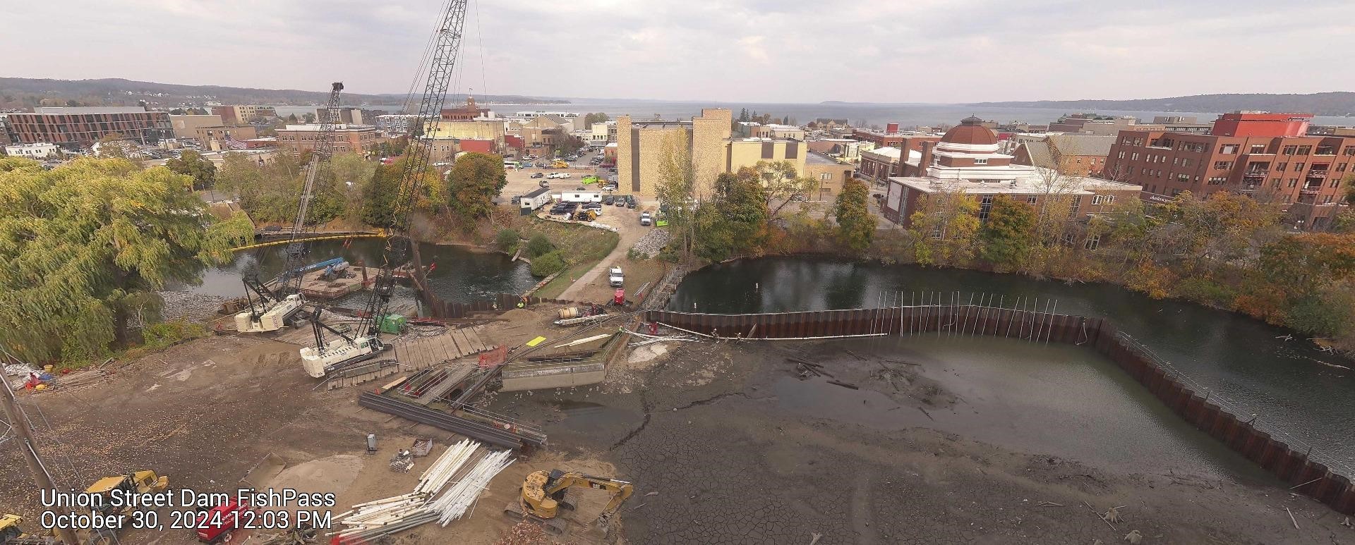 Aerial view of the Union Street Dam FishPass construction site, taken on October 30, 2024, at 12:03 PM. The site shows significant progress, with heavy equipment, including excavators and cranes, visible on the construction area. Sheet piling sections divide parts of the river, and exposed riverbed sections are visible due to water diversion. In the background, urban buildings, trees with autumn foliage, and a view of the bay under an overcast sky are visible.